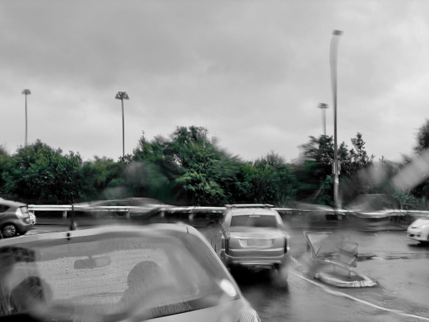 Busy intersection filled with cars in a rainy day. Only the trees from afar are coloured in green. Everything else is gray.